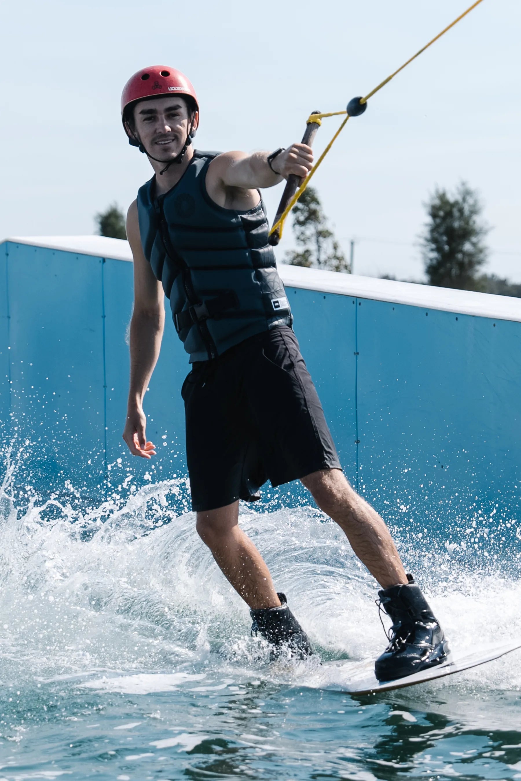 Person wakeboarding on a body of water with a blue wall in the background, wearing a Jetpilot X1 Life Jacket - Bust Dunn Signature Series - Charcoal