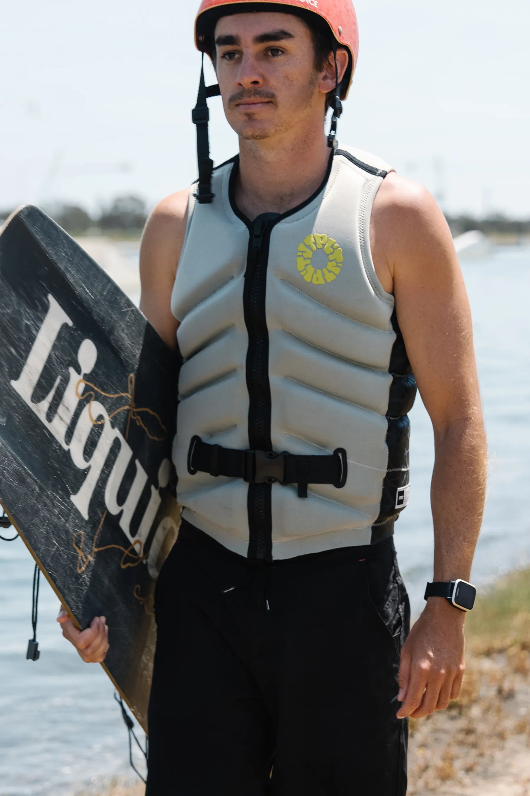Person wearing a Jetpilot X1 Life Jacket - Busty Dunn Signature Series - Light Grey holding a wakeboard branding near water.