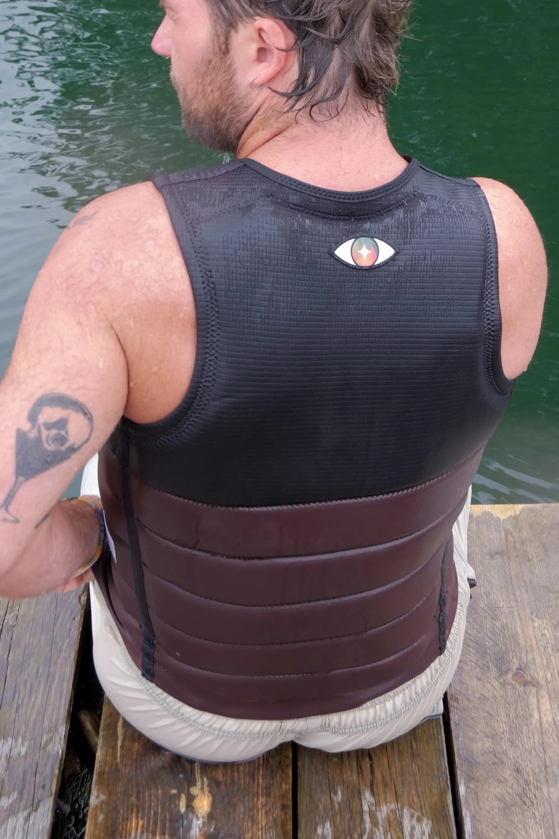 Man wearing a black and brown life vest sitting on a wooden dock by water.