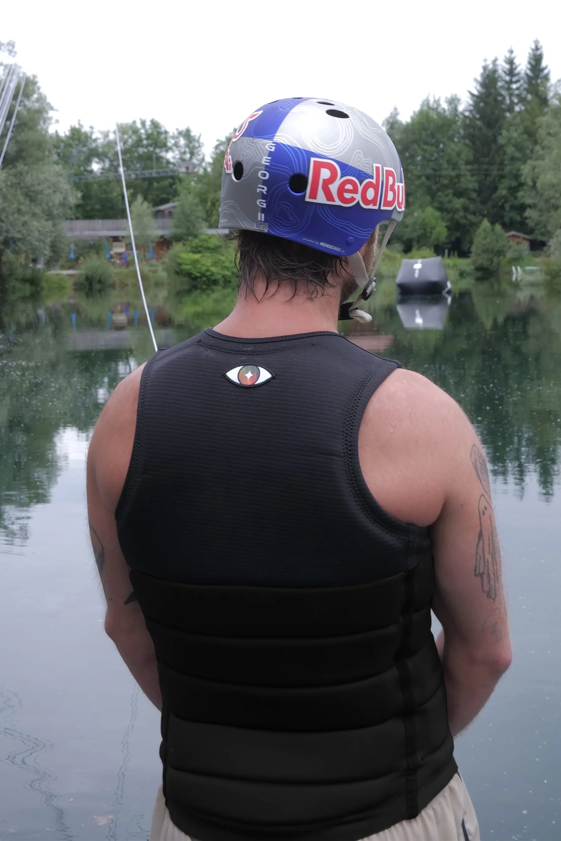 Person wearing a Jetpilot X1 Life Jacket - Felix Georgii Signature Series - Black and RedBull helmet by a lake