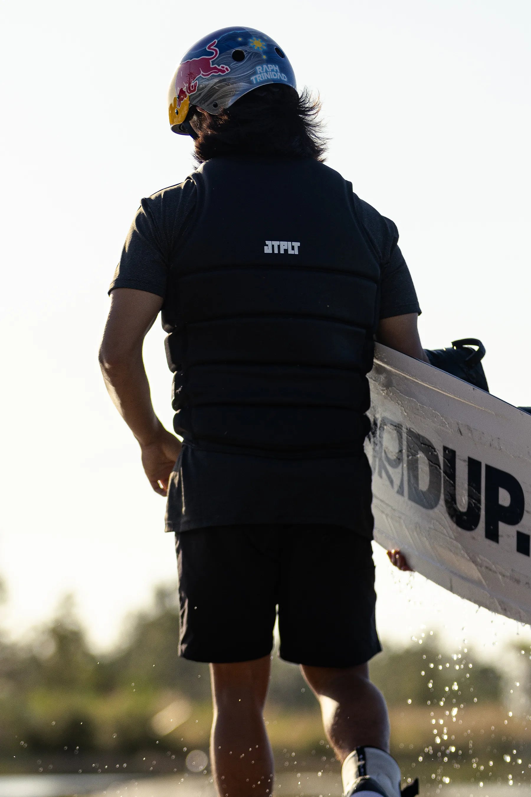 Person holding a wakeboard with a helmet on, in a Jetpilot X1 Life Jacket - Raph Trinidad Signature Series  - Black standing on a beach.