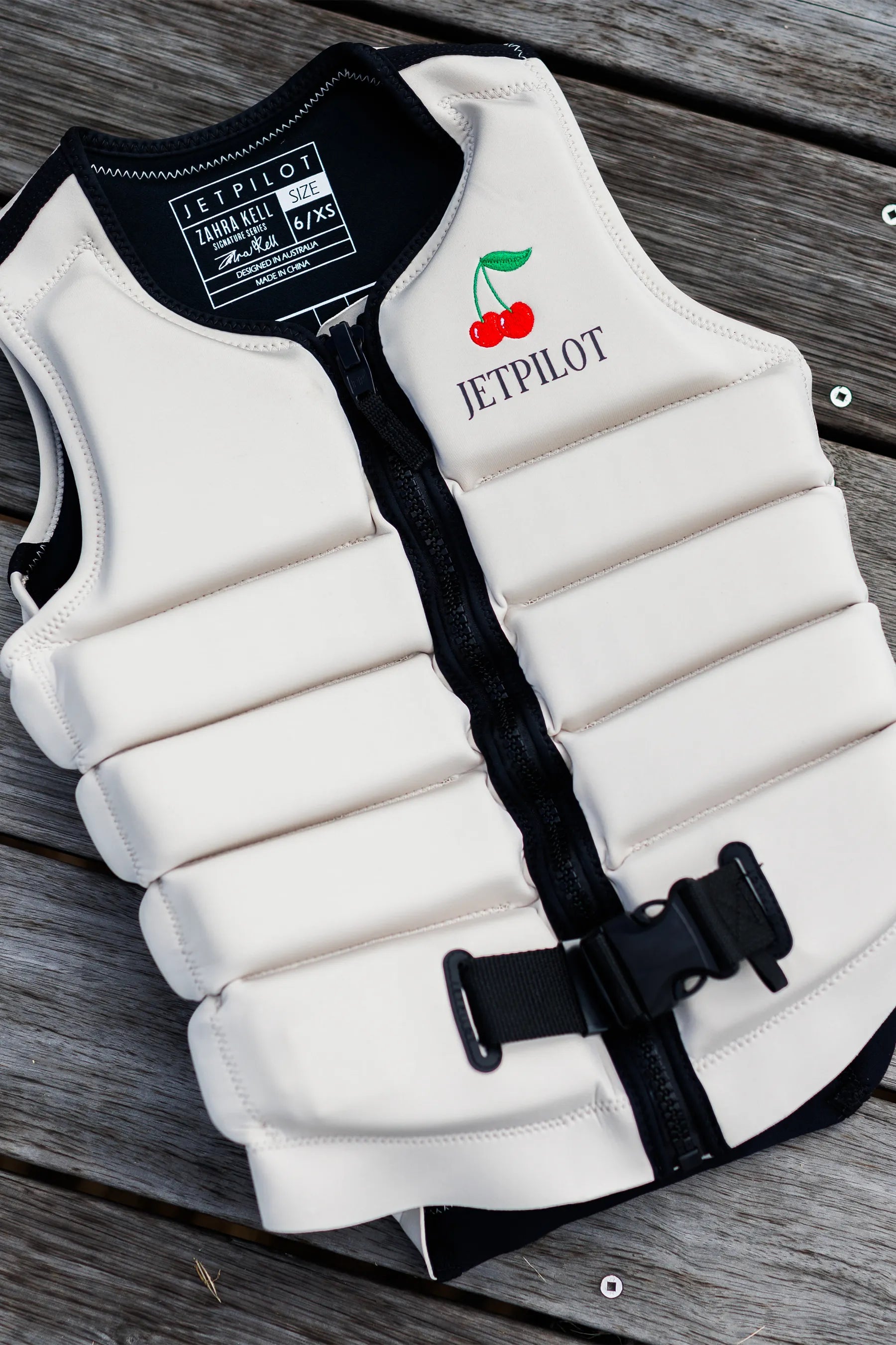 White Life jacket with black straps and Jetpilot logo, embroidered red cherries on a wooden surface