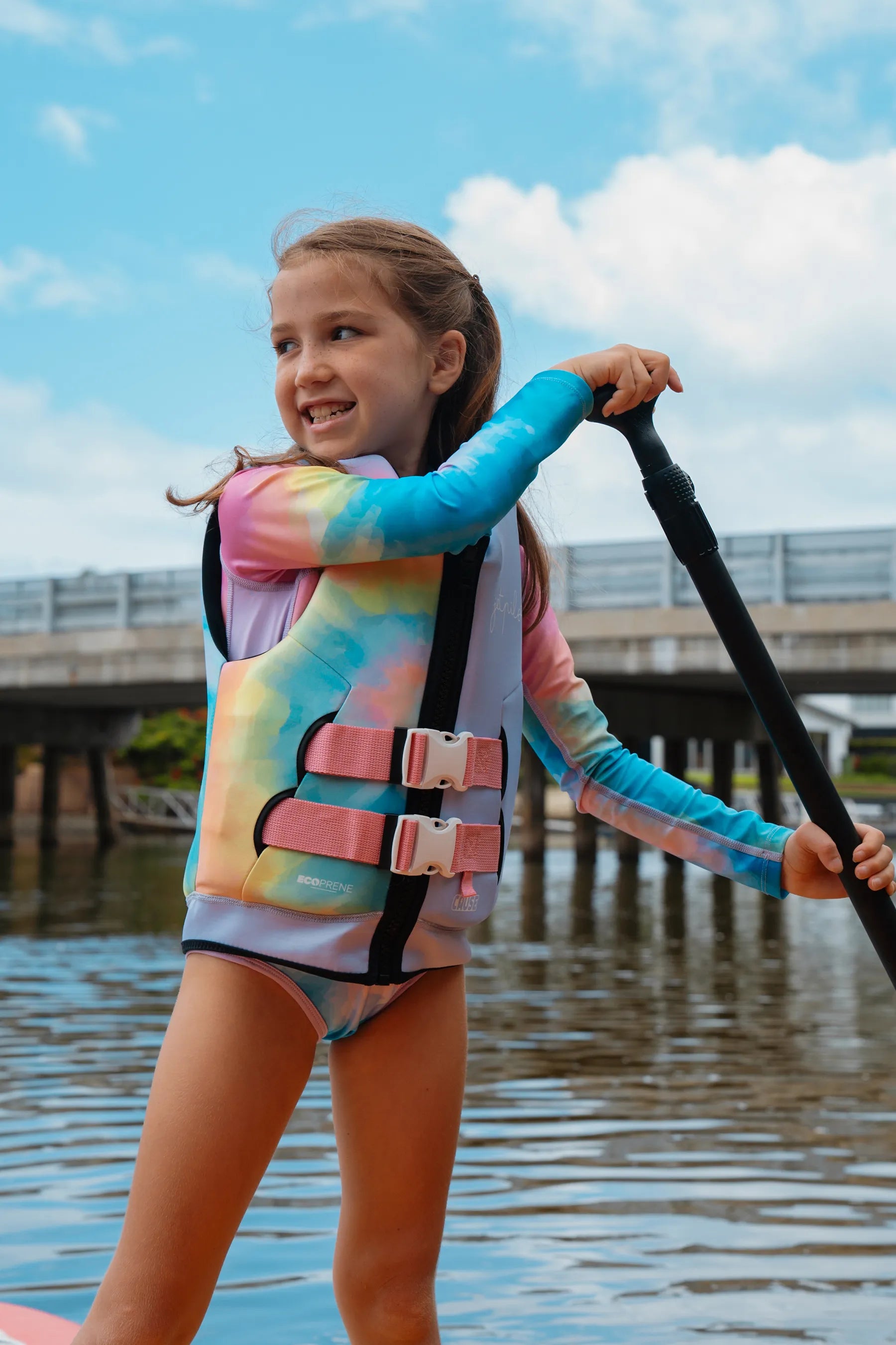 Child wearing a colorful life jacket and holding a paddle on a body of water.