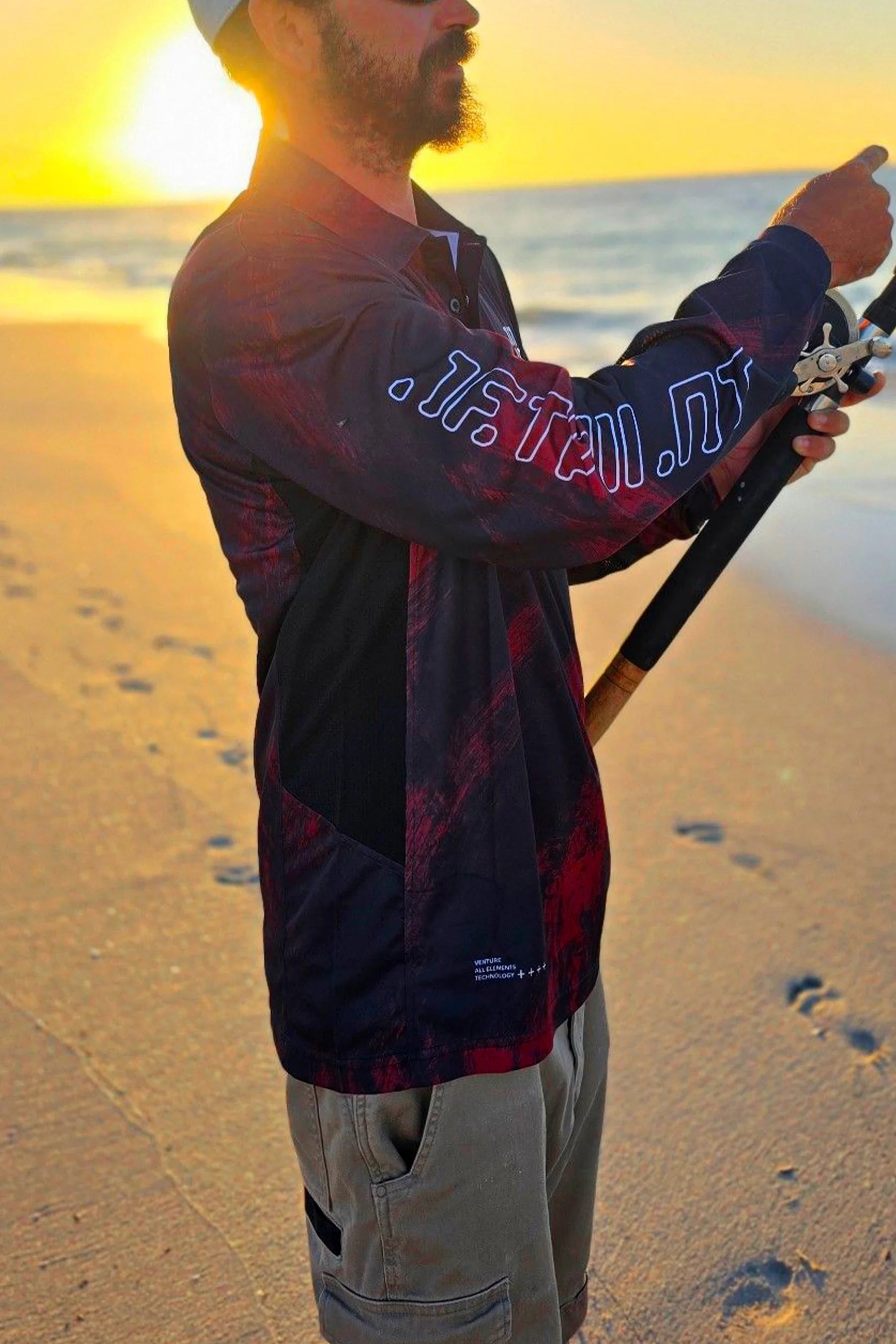 Person on a beach holding a fishing rod with a sunset in the background wearing the Jetpilot Venture Long Sleeve Fishing Polo - Red