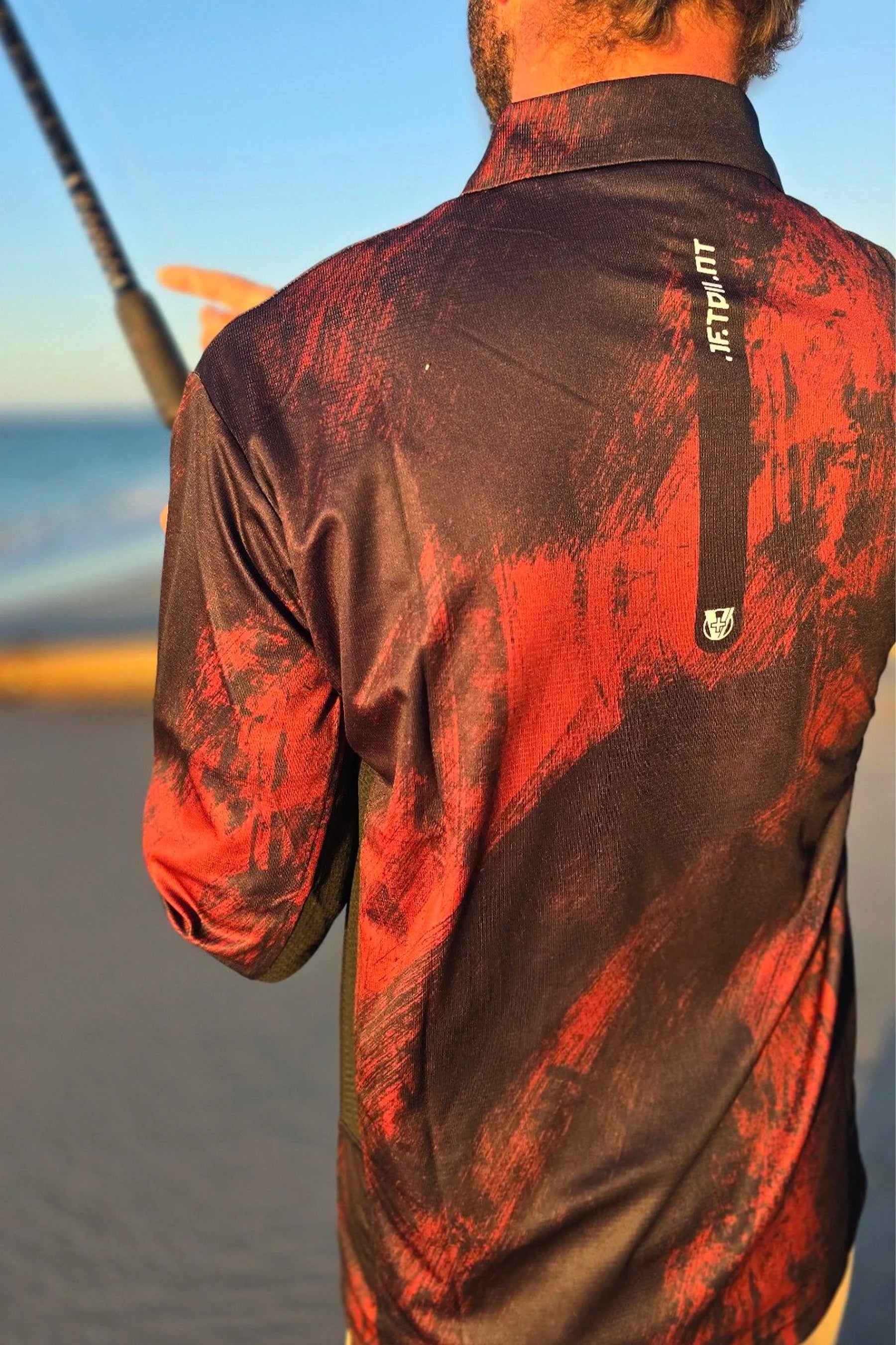 Person wearing Jetpilot Venture Long Sleeve Fishing Polo - Red and black patterned shirt with a logo on a beach.
