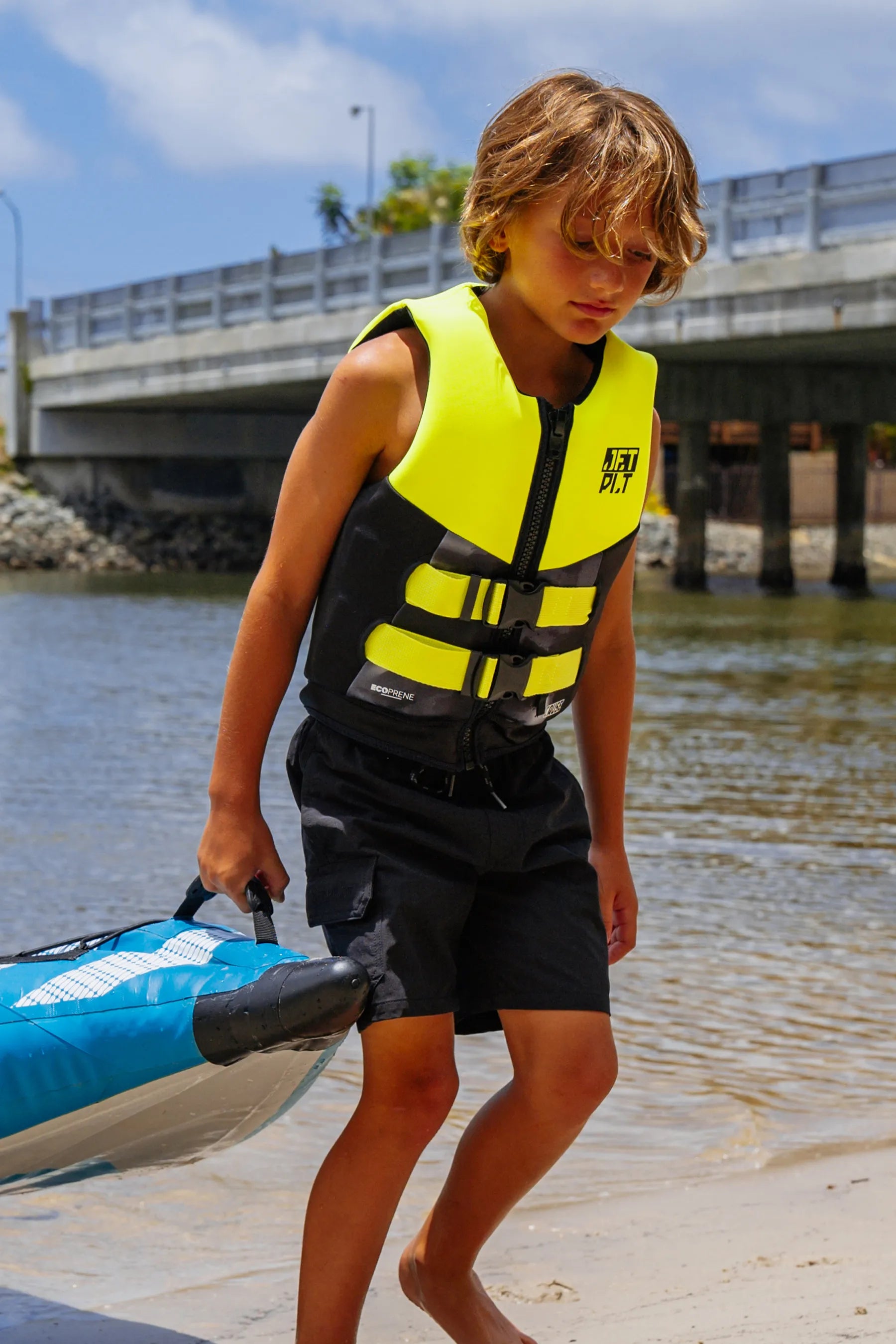 Child wearing a Cause Kids Life Jacket- Yellow L50 holding a kayak by a waterfront with a bridge in the background.