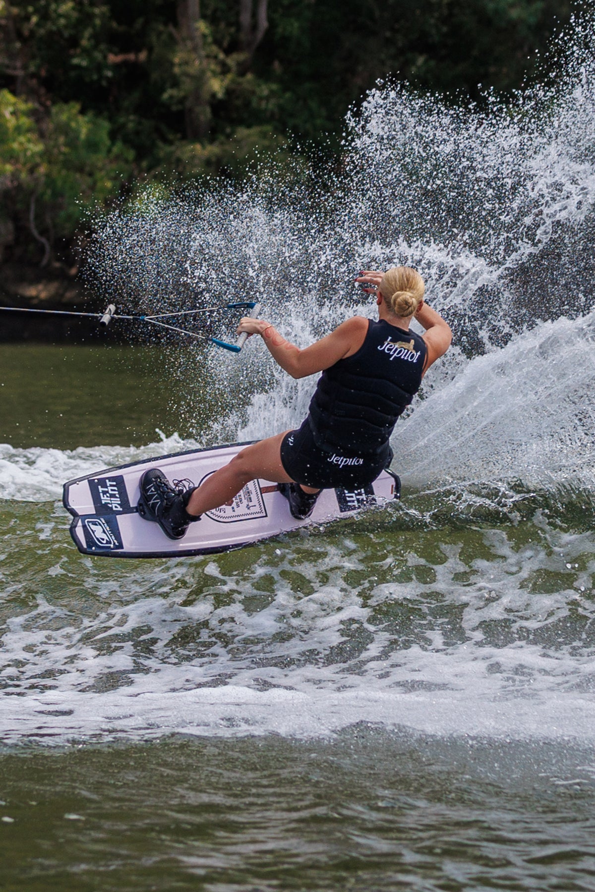 Rider carving on a wakeboard behind the boat and throwing spray, wearing the Black Zahra X1 life jacket